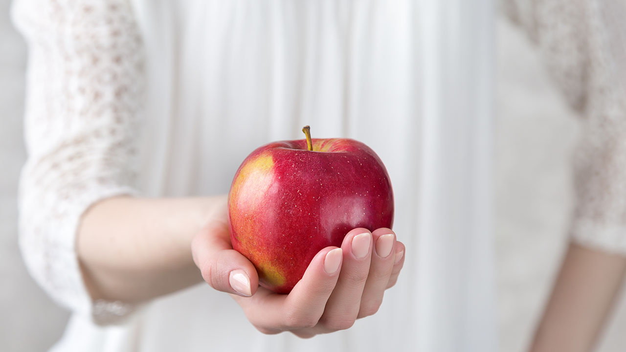 A person wearing a white lace sleeve holds a red apple in their outstretched hand, offering a simple health hack for daily wellness.