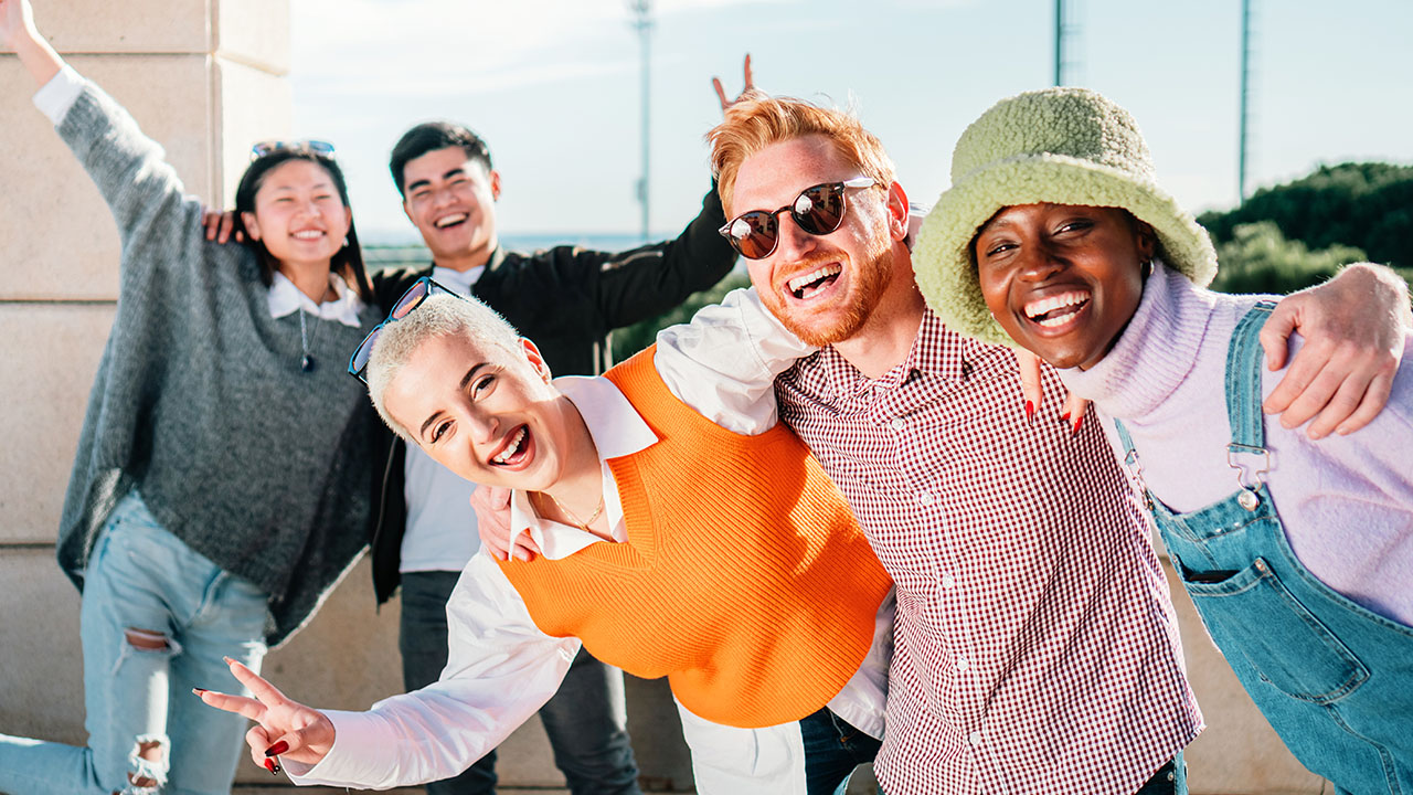 A group of five people stands outdoors on a sunny day, smiling and posing together with some making peace signs, as if celebrating their latest health hacks.