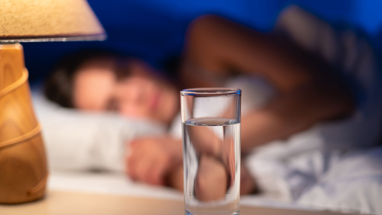 A glass of water on a bedside table with a person sleeping peacefully in the background illustrates one of the simplest health hacks for staying hydrated overnight.