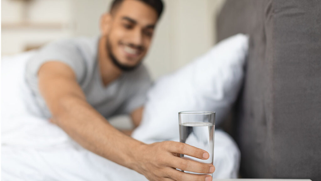 A man in a gray shirt reaches out to pick up a glass of water from a bedside table while sitting in bed, a simple health hack to stay hydrated throughout the night.