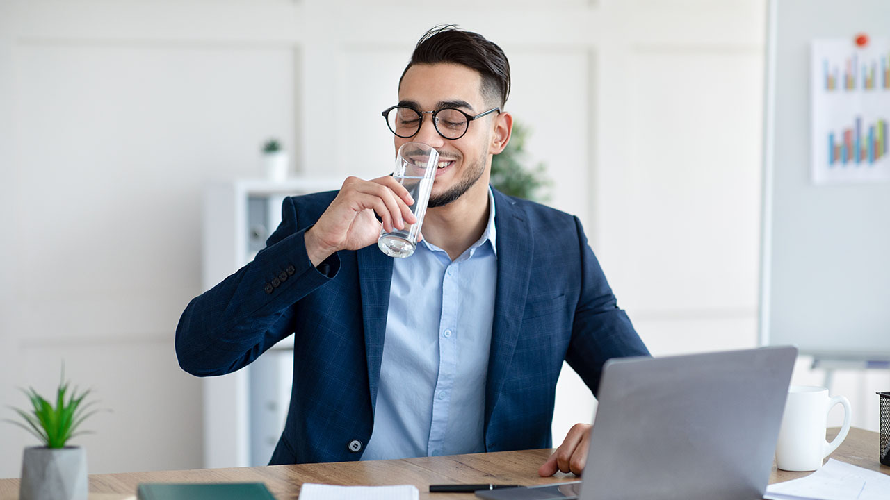 A man in a blue suit and glasses is sitting at a desk with a laptop, holding and drinking from a glass of water—a simple Health Hack—while smiling.