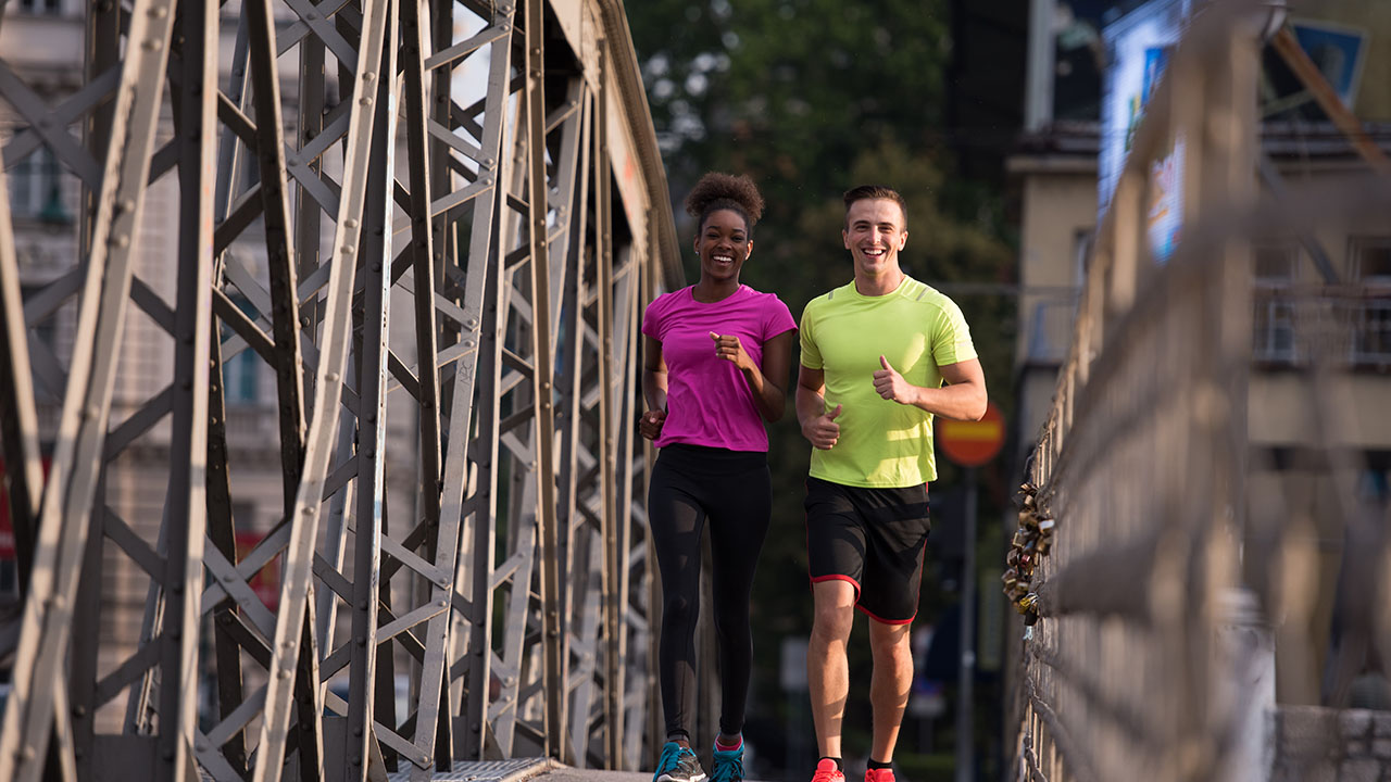 Two people jog on a bridge, both wearing brightly colored athletic clothing. The person on the left is in a pink shirt and black leggings, and the person on the right is in a yellow shirt and black shorts. Their routine embodies simple health hacks for staying fit and energized.