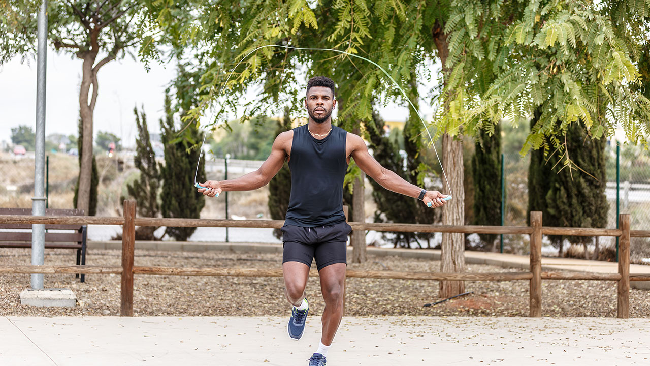 A man in athletic wear expertly jumps rope in an outdoor park setting, with trees and a wooden fence in the background—a perfect example of health hacks in action.