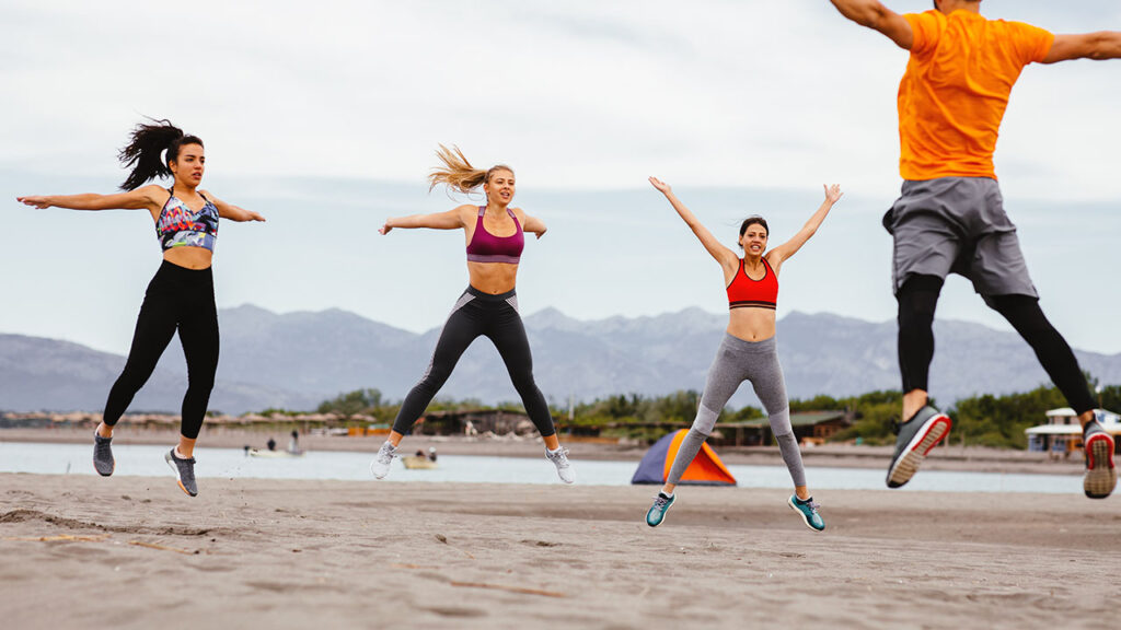 Four people perform jumping exercises on a sandy beach, with mountains and tents visible in the background, showcasing one of nature's best health hacks.