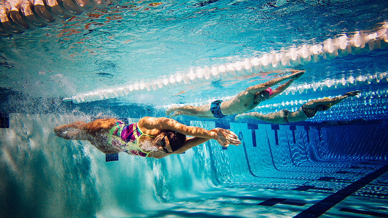 Two swimmers in a pool are captured underwater as they swim side by side, with one in a pink suit and the other in a blue suit, both performing the front crawl stroke—a perfect example of fitness and health hacks.