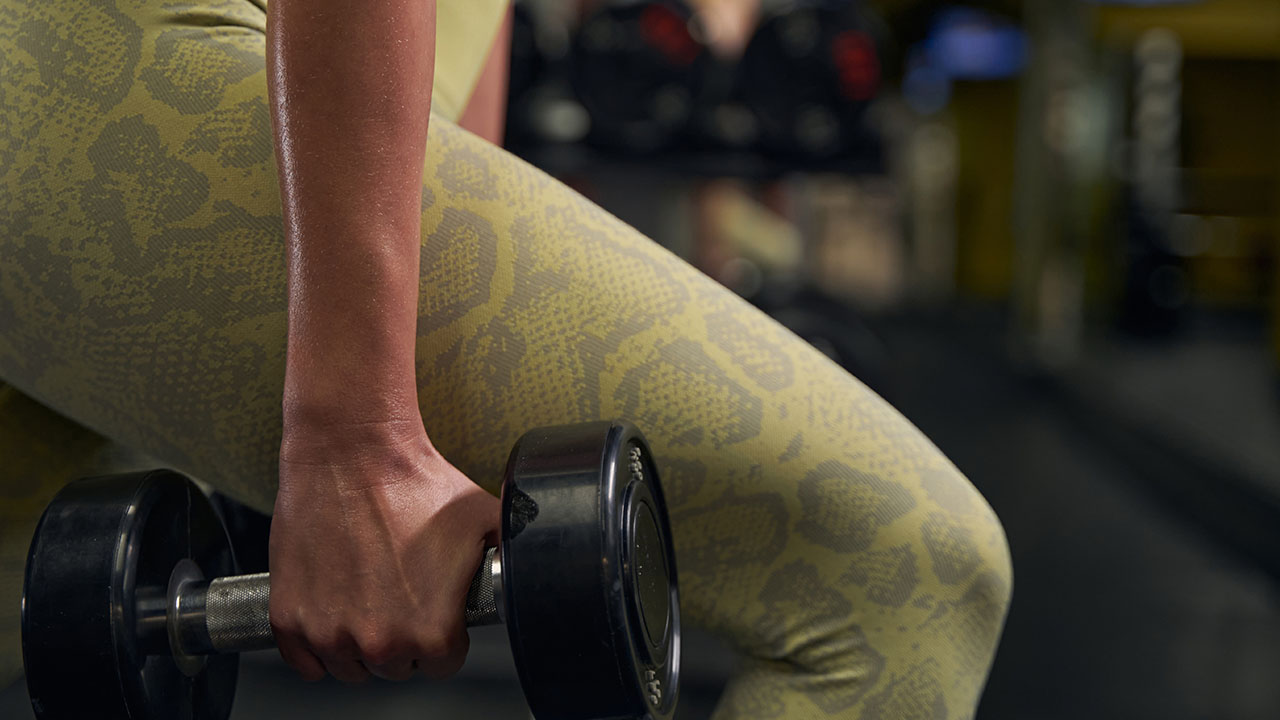 Close-up of a person wearing yellow leggings holding a dumbbell in a gym. Background is blurred with gym equipment visible, embodying the essence of health hacks for fitness enthusiasts.