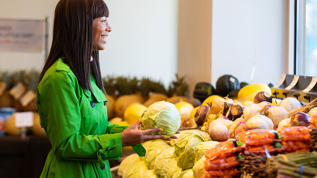 Woman in a green coat holds a cabbage, exploring health hacks while shopping for vegetables in a grocery store.