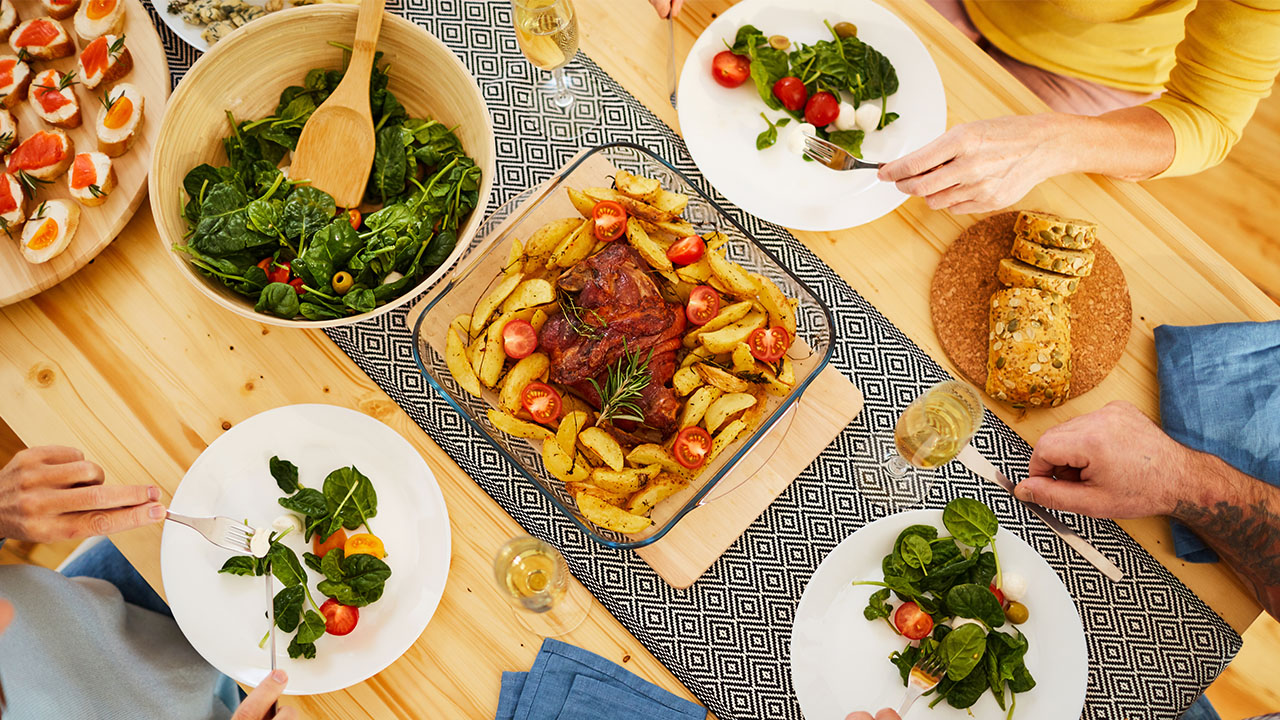 A table setting with a baked dish of meat and potatoes, salad bowls, bread, and plates of salad. Three people are seated, holding forks. Blue napkins and glasses of white wine add a touch of elegance to this meal inspired by health hacks for balanced eating.