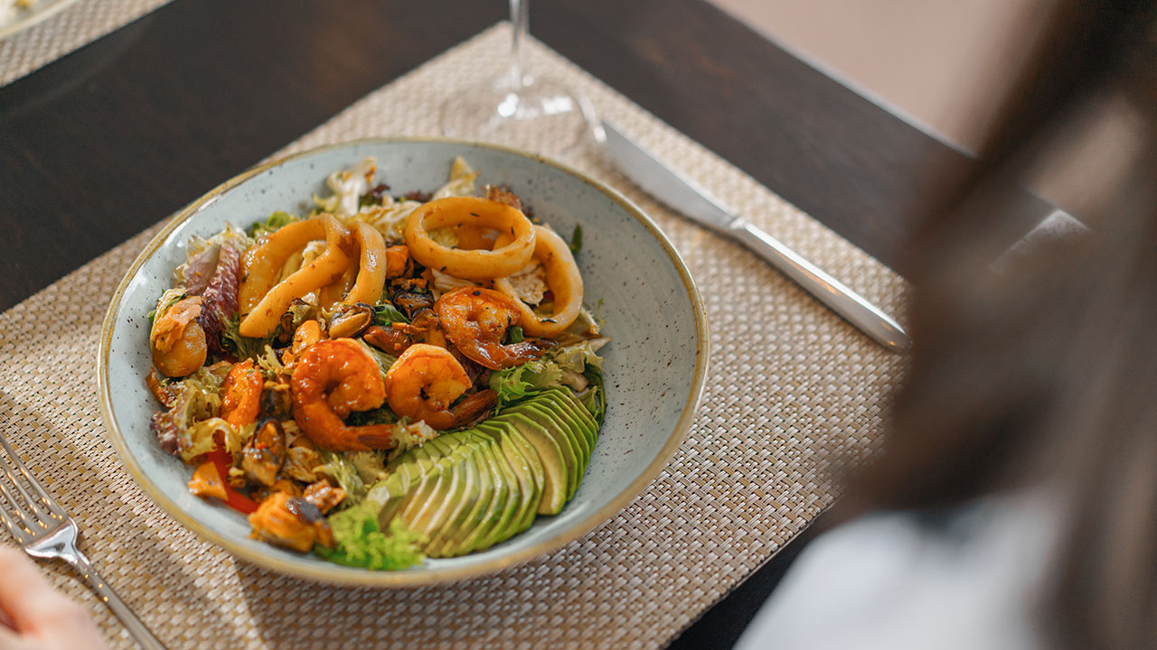 A bowl of salad featuring shrimp, calamari, avocado slices, and an assortment of vegetables is placed on a woven placemat, with a person sitting at the table enjoying health hacks in every bite.