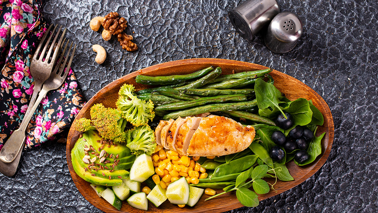 A wooden bowl filled with grilled chicken, green beans, broccoli, avocado, cucumber, corn, spinach, and blueberries sits next to a colorful cloth napkin. Cutlery, nuts, and salt and pepper shakers complete this delightful meal that's perfect for your health hacks arsenal.