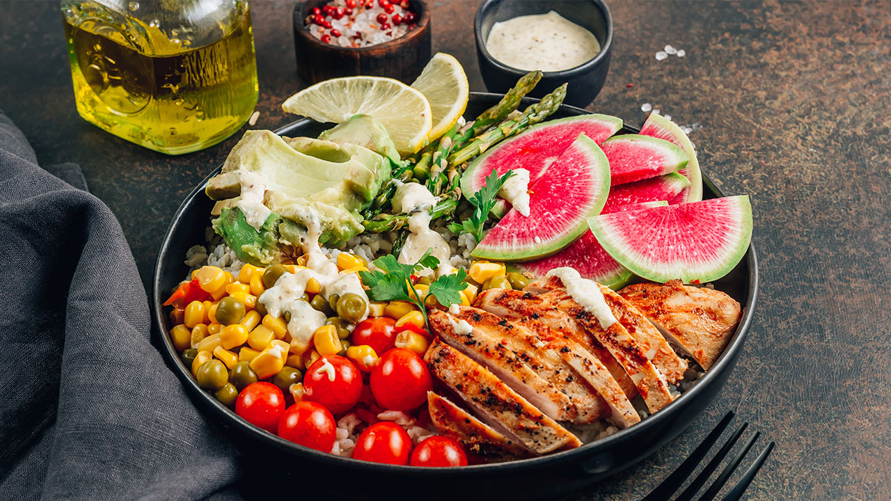 A colorful bowl featuring grilled chicken, cherry tomatoes, corn, peas, asparagus, sliced watermelon radish, avocado, and lemon slices, garnished with dressing. Oil and condiments are in the background—a perfect meal for those seeking health hacks.