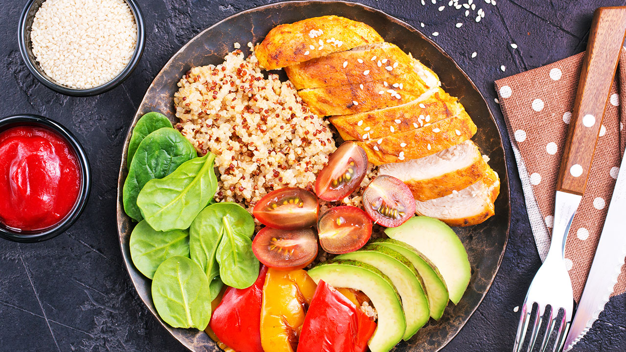 A bowl with grilled chicken, quinoa, spinach, cherry tomatoes, avocado slices, and bell peppers—your ultimate health hack. Fork and knife are elegantly placed on a polka dot napkin next to the bowl. A side of sauce and sesame seeds completes this nutritious meal.