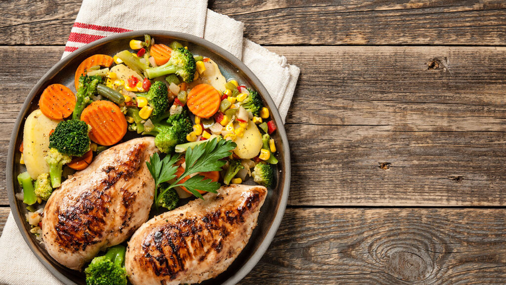 A plate with two grilled chicken breasts and a mix of vegetables, including broccoli, carrots, corn, and potato slices, sits on a wooden table with a striped cloth napkin beside it—a perfect meal for those exploring Health Hacks.