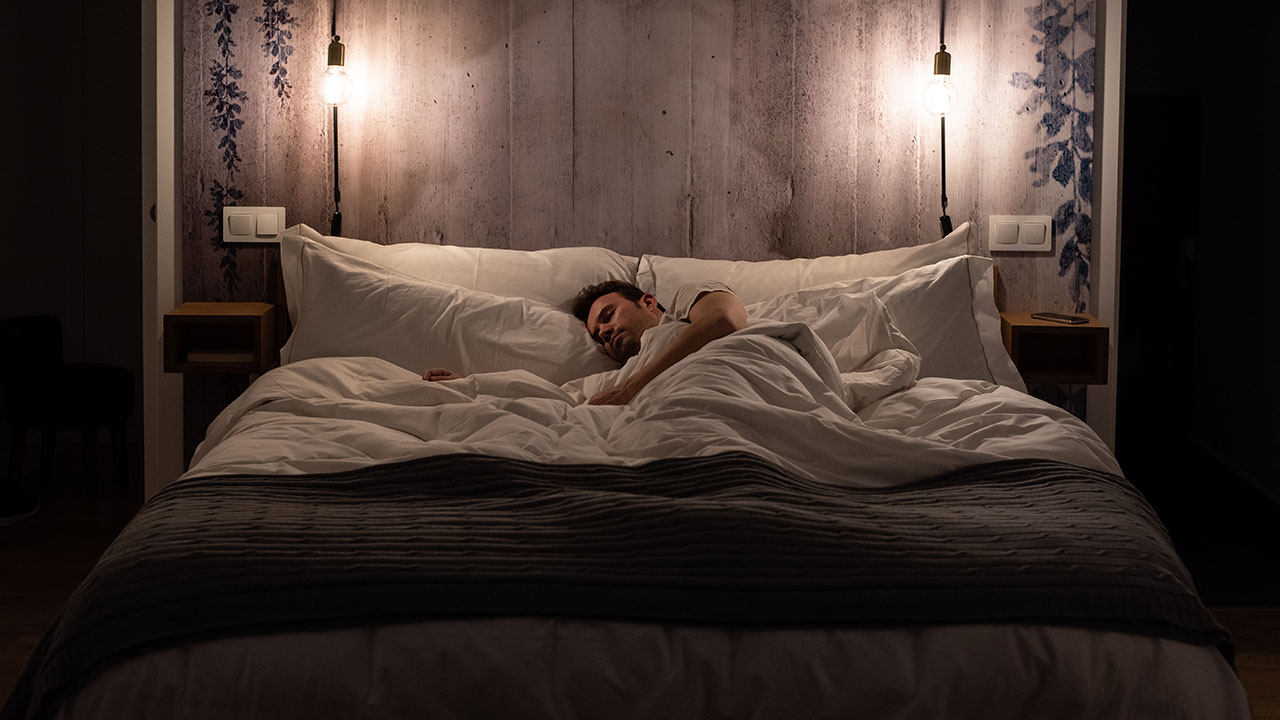 A person sleeps on a neatly made bed with white bedding and two illuminated bedside lamps, embracing one of the best health hacks for better rest, all against a wooden backdrop.