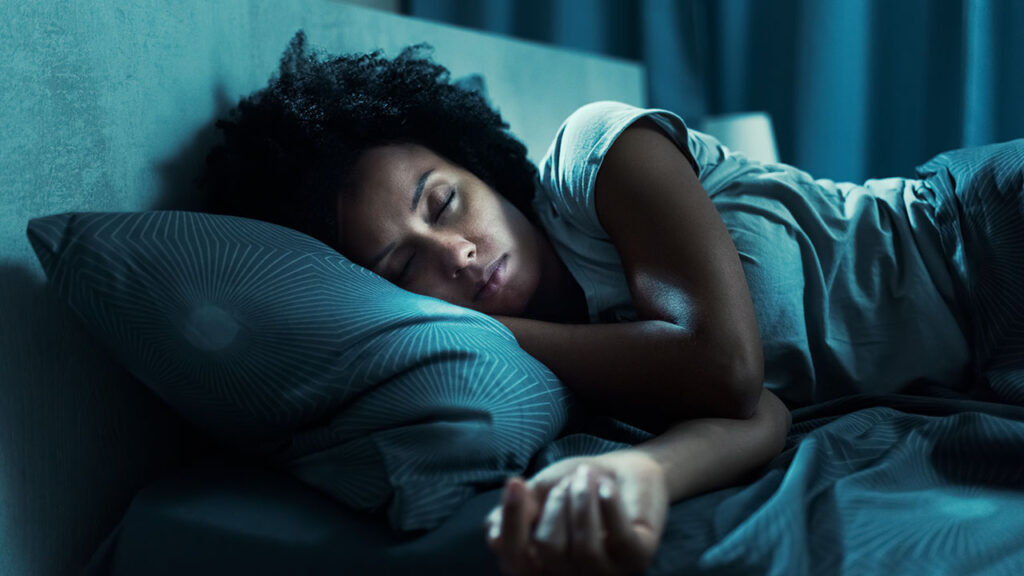 A person with curly hair lies peacefully on a bed, resting their head on a patterned pillow and covered with a similar blanket. The room is softly lit, suggesting nighttime—a perfect setting for practicing some calming health hacks before sleep.