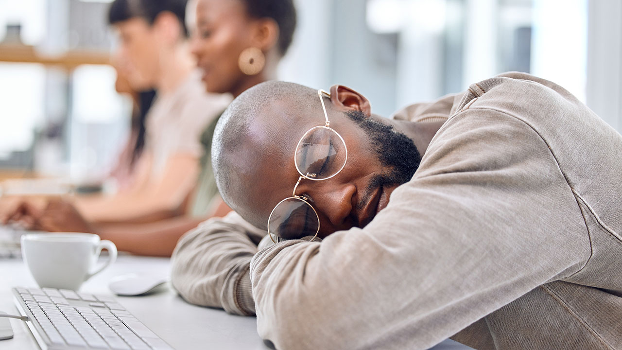 A person wearing glasses and casual clothing is asleep on a desk in an office setting, with two other people working in the background, possibly pondering some health hacks to boost their energy levels.