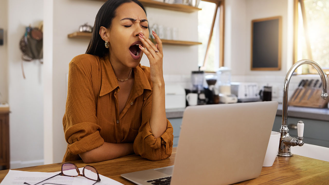 A person in a brown shirt yawns while sitting at a kitchen counter with an open laptop and glasses, perhaps researching some health hacks.