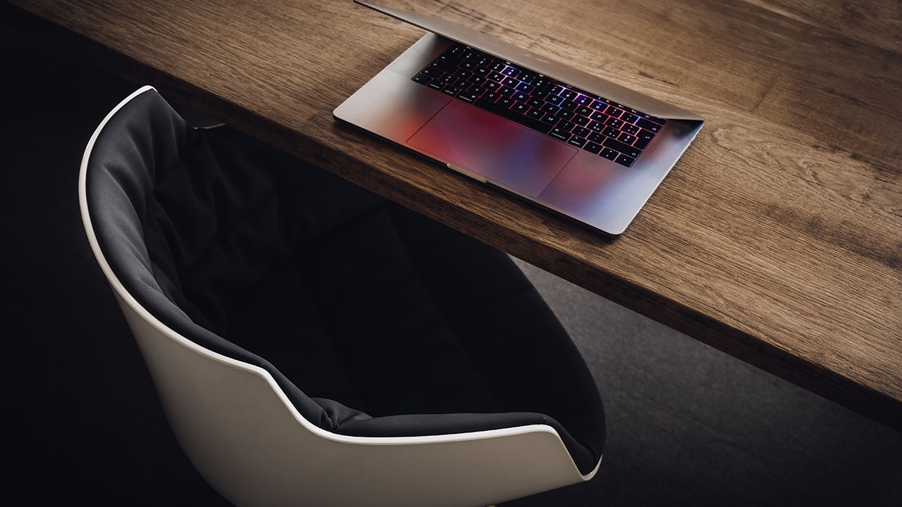 A laptop with a partially closed lid sits on a wooden desk next to a black cushioned chair, embodying the perfect setup for exploring the latest health hacks.