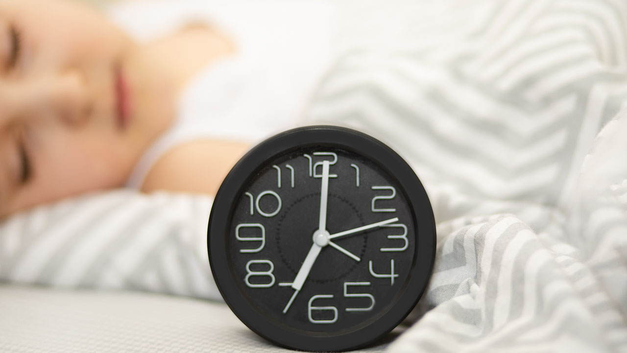 A child is sleeping under a gray and white striped blanket with a black analog clock showing 3:55 in the foreground, demonstrating one of the health hacks for maintaining a restful night's sleep.