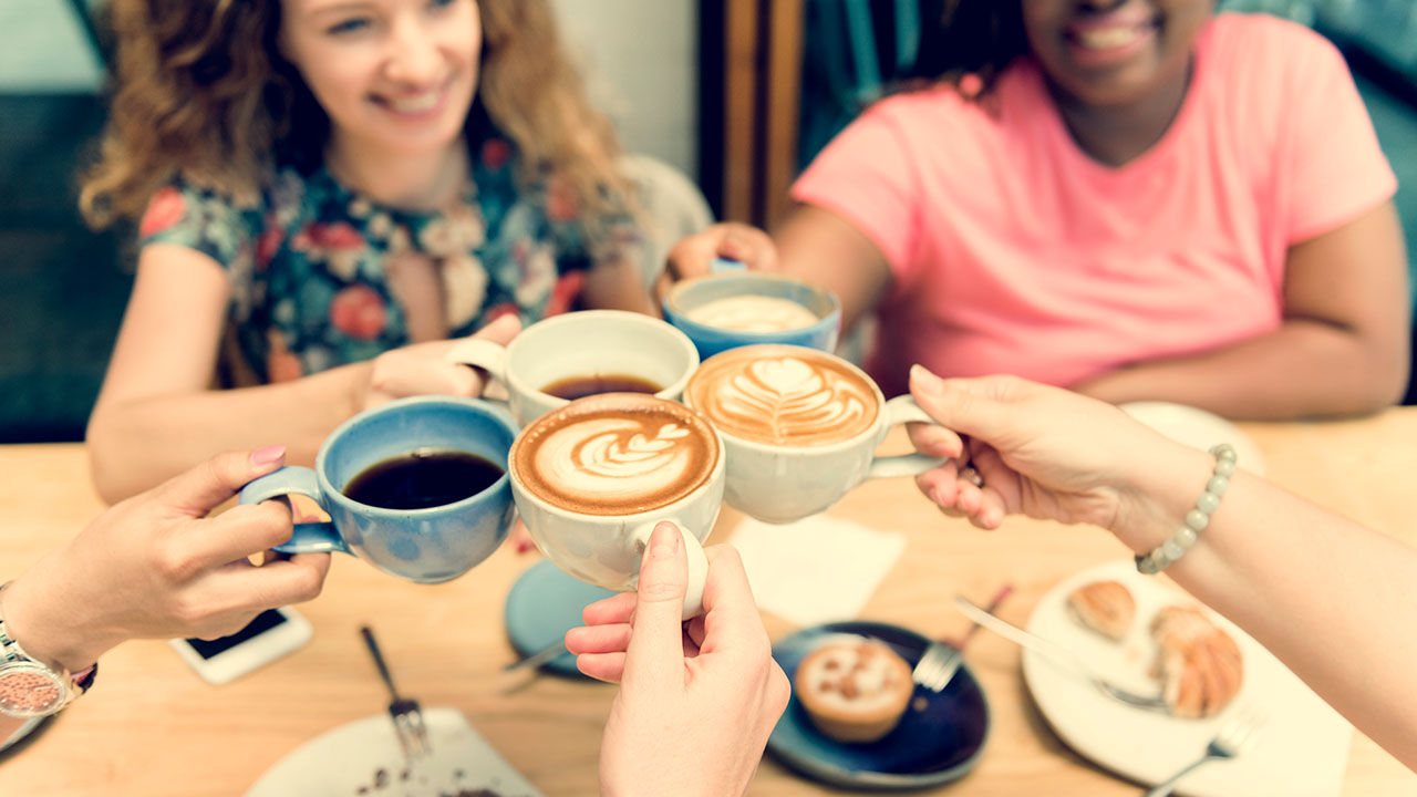 Four people clink coffee mugs together over a table with plates of food, sharing not just a meal but also their favorite health hacks.