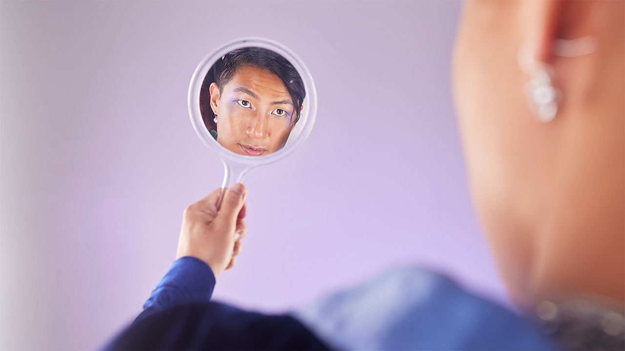 A person is holding a round handheld mirror, looking at their reflection. With health hacks in mind, they sport a focused expression as the blurred background highlights their intent gaze.