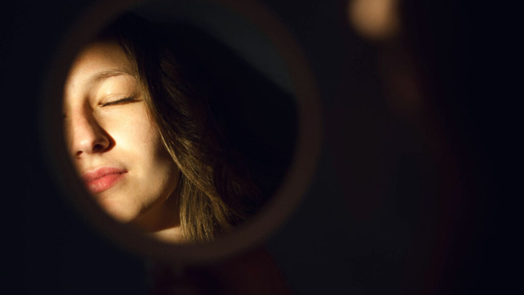 A person's face is partially illuminated while reflected in a round mirror, with one eye closed and lips slightly upturned, as if contemplating health hacks.