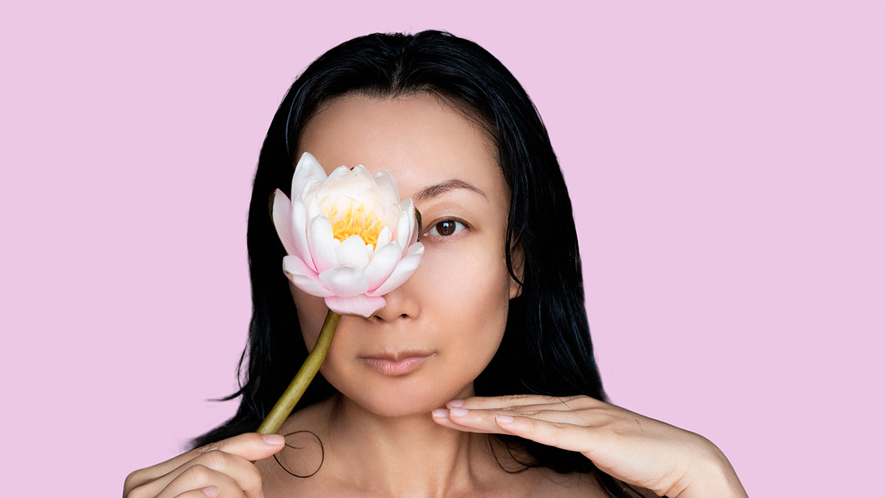 A woman with long dark hair holds a pink and white lotus flower in front of her left eye against a plain pink background, embodying the essence of health hacks. Her right hand is placed near her chin.