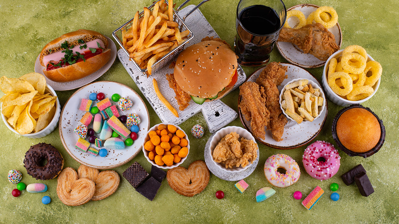 A variety of fast food and snacks including a burger, fries, hot dog, fried chicken, donuts, candy, and a drink displayed on a green background. Consider these health hacks to enjoy your favorites in moderation!