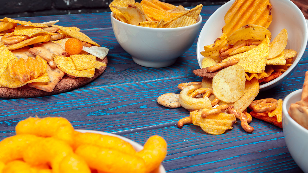 Various bowls and plates filled with assorted snacks, including potato chips, cheese puffs, and pretzels, on a blue wooden table. Some snacks have spilled onto the table—an inviting scene for a cozy gathering. For those mindful of health hacks, adding veggie sticks or nuts can balance this spread delightfully.