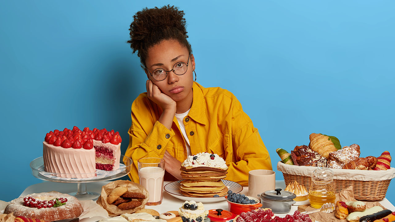 A person wearing glasses and a yellow shirt sits at a table with various desserts, including a strawberry cake, pancakes, pastries, and a glass of milk, enjoying the sweet indulgence while pondering health hacks against a blue background.