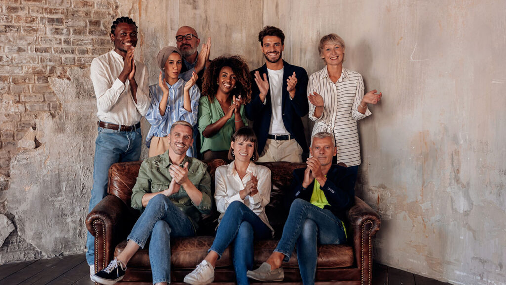 A diverse group of eight people, standing and sitting on a couch, are clapping and smiling in a rustic, exposed brick indoor setting, celebrating their latest health hacks together.