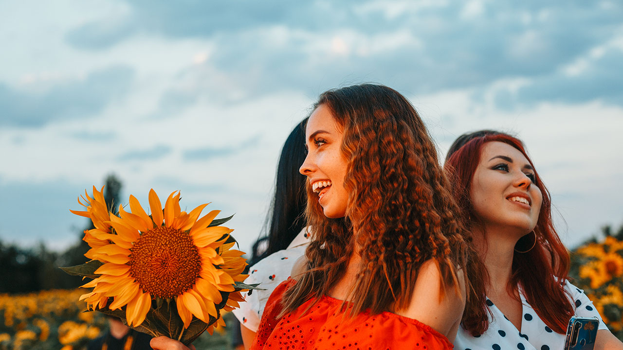 Three women smile and enjoy a sunny day in a sunflower field, with one woman holding a large sunflower. The vibrant scene radiates happiness, proving that nature's simple pleasures are among the best health hacks around.