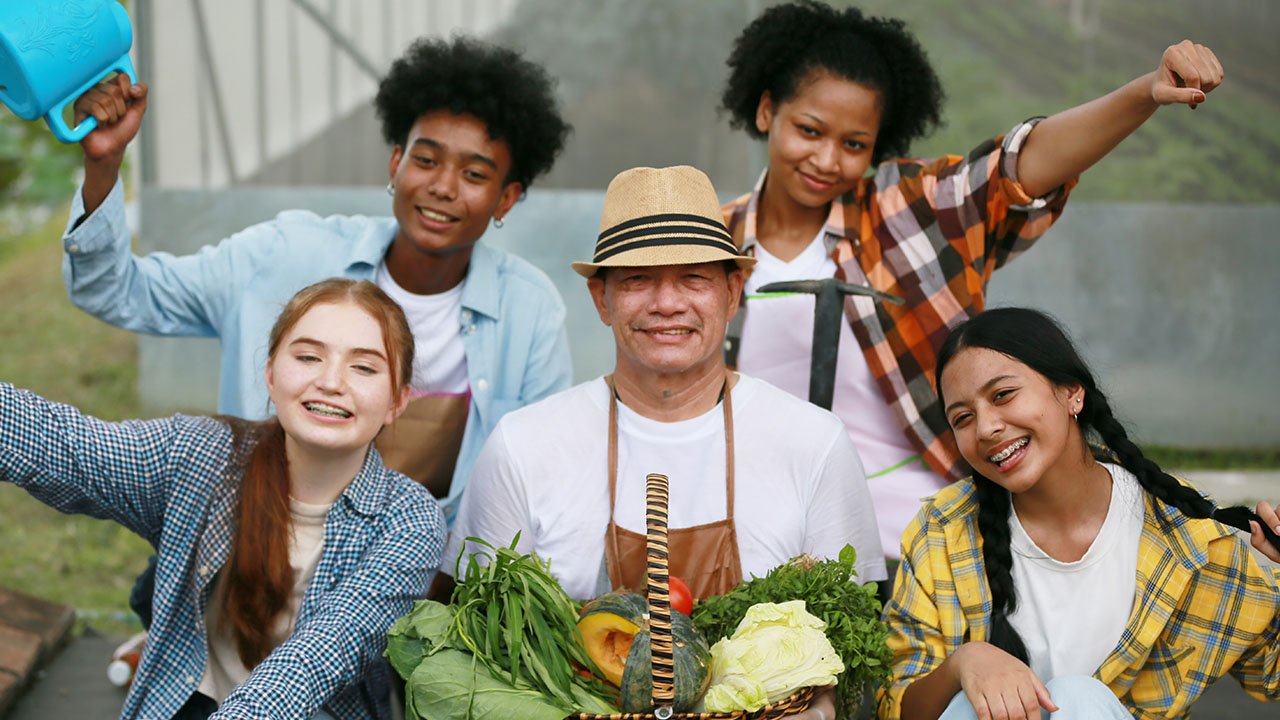 A group of five people, including a middle-aged man with a straw hat holding a basket of vegetables, smiles and poses together outdoors, embodying simple health hacks. Two young women and two young men surround him, radiating joy and vitality.