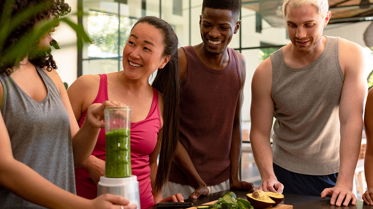 Four people at a kitchen counter watch as one person uses a blender filled with green smoothie ingredients. They are smiling and appear engaged in the activity, exploring some fun health hacks together.