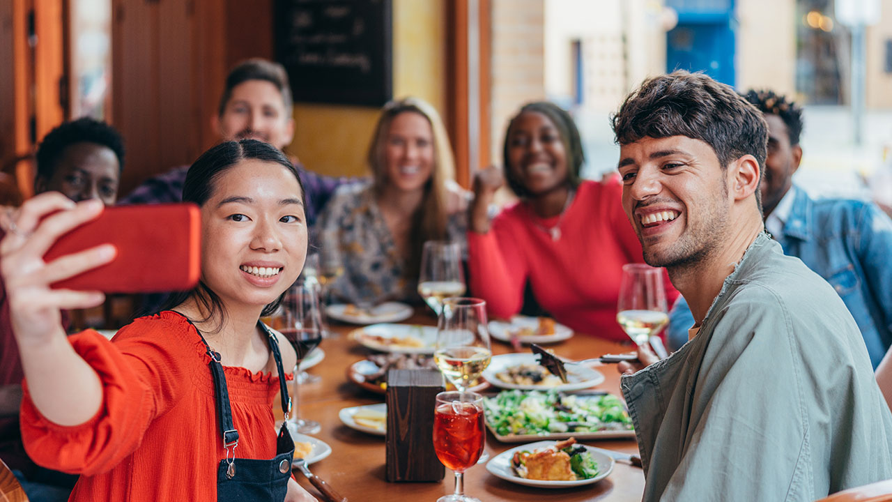 A group of people sitting at a restaurant table, smiling and taking a selfie. Plates of food featuring some clever health hacks and glasses of drinks are on the table.