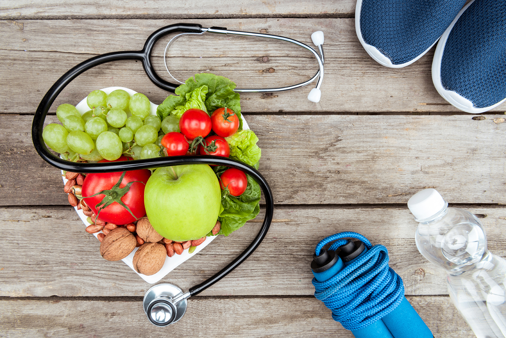 A stethoscope encircles a heart-shaped bowl filled with fruits and vegetables on a wooden surface, flanked by athletic shoes, a jump rope, and a water bottle—perfect for highlighting health themes in your SEO strategy.