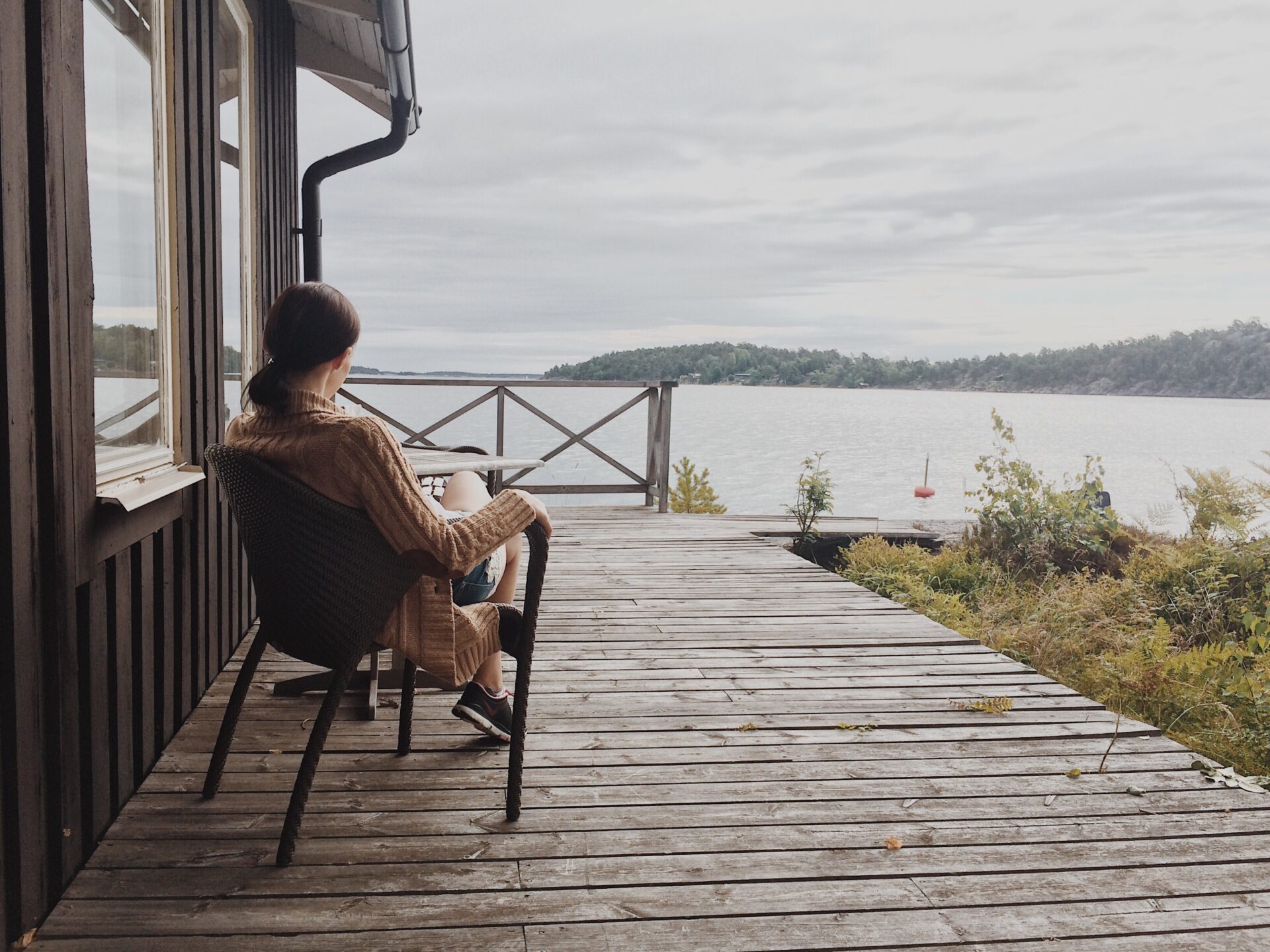 A woman sitting in a wicker chair on a wooden deck overlooking a lake with overcast skies.