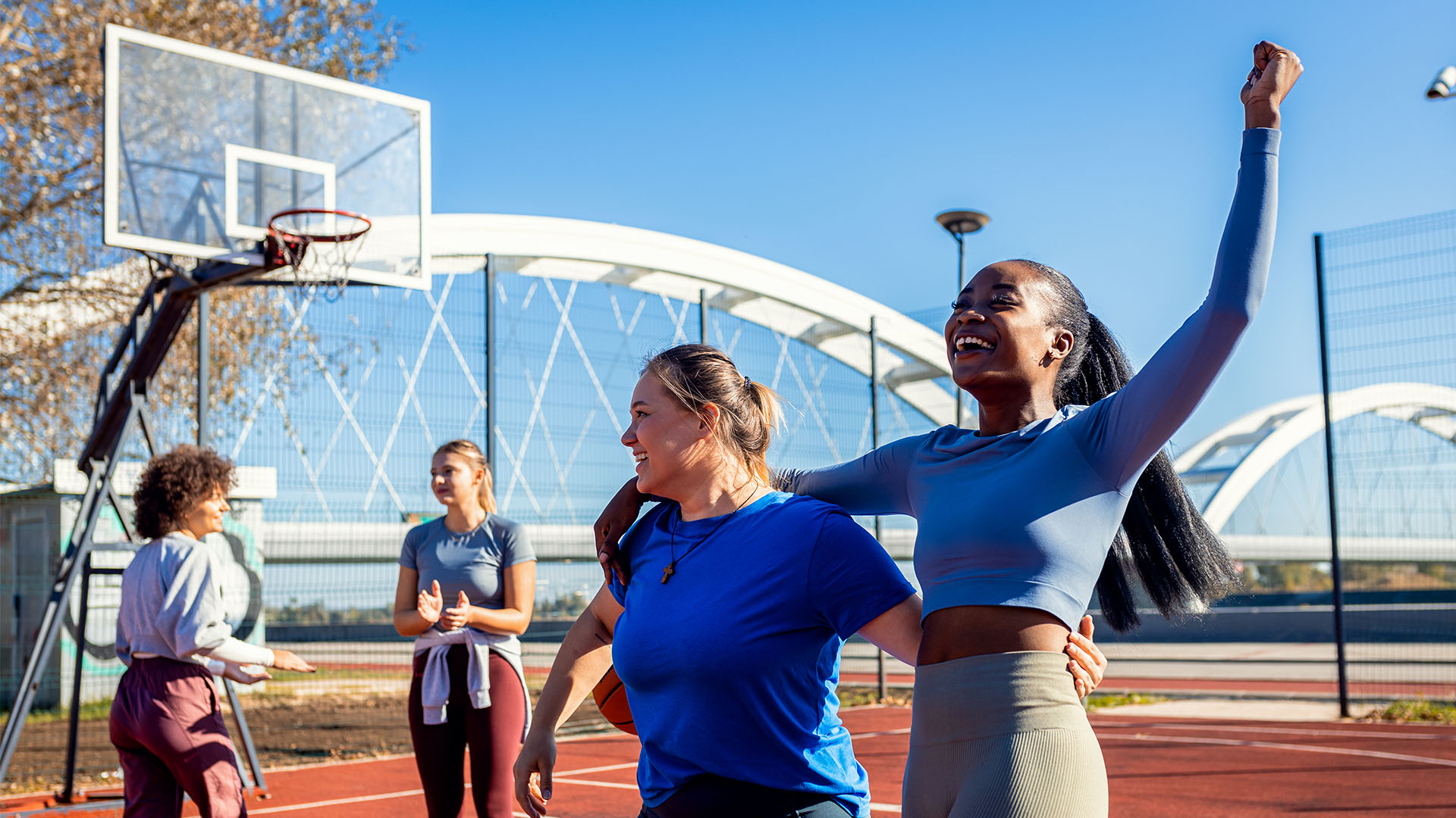 Four women gather on an outdoor basketball court. Two women in the foreground are smiling and celebrating, embodying the perfect gym camaraderie, while two others in the background are clapping and cheering.