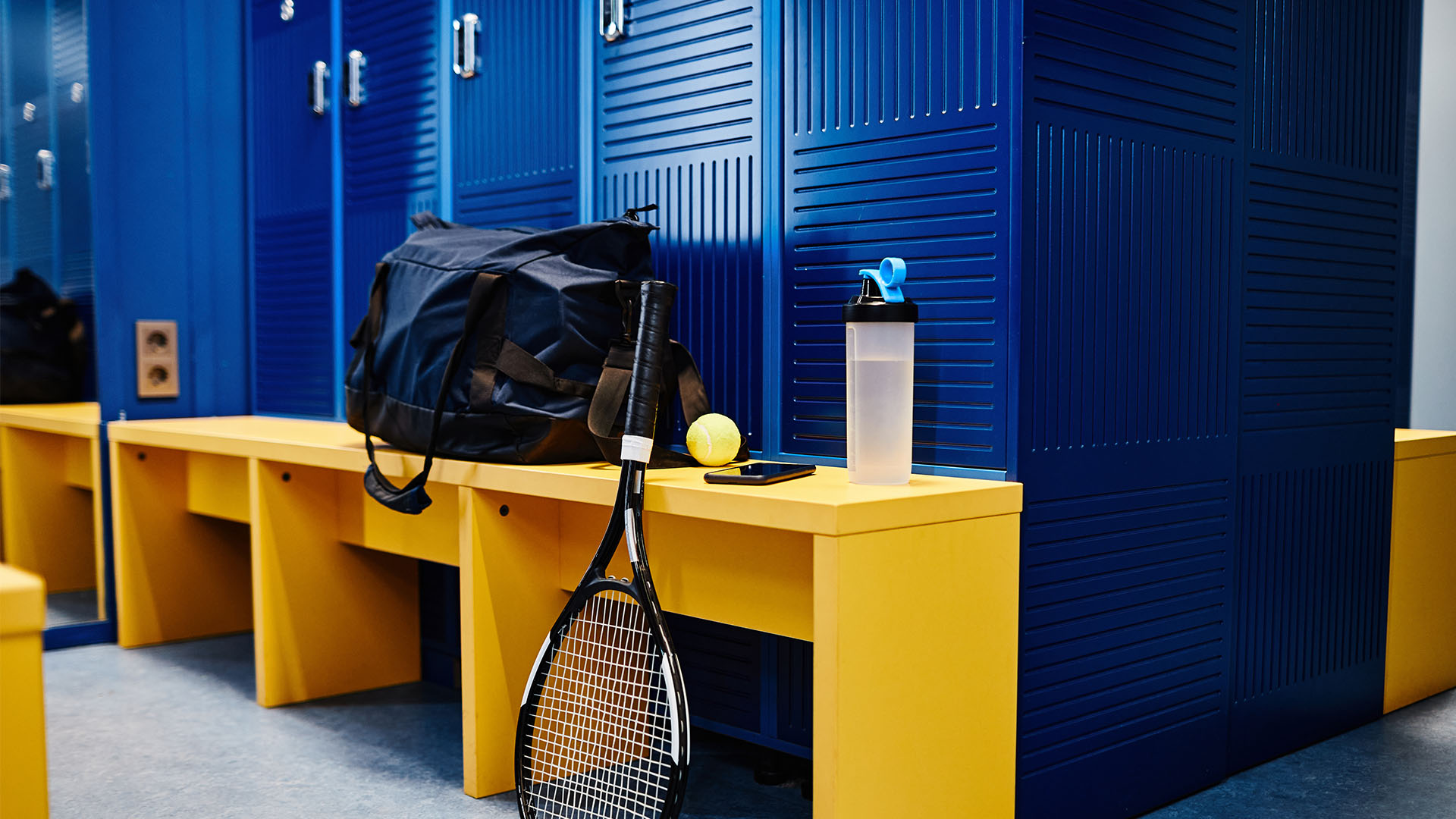 A gym locker room with blue lockers and yellow benches. A duffel bag, tennis racket, ball, water bottle, and phone are placed on a bench. If you’re looking for gym tips, always keep your essentials organized to avoid wasting time searching for them.