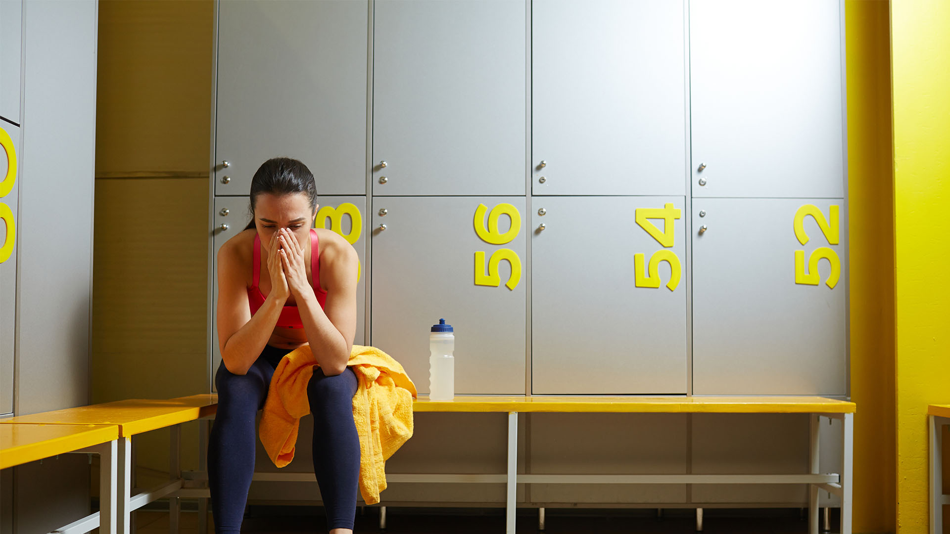 A person in workout attire sits on a bench in a gym locker room, looking down with hands covering their face, next to a yellow towel and a water bottle.