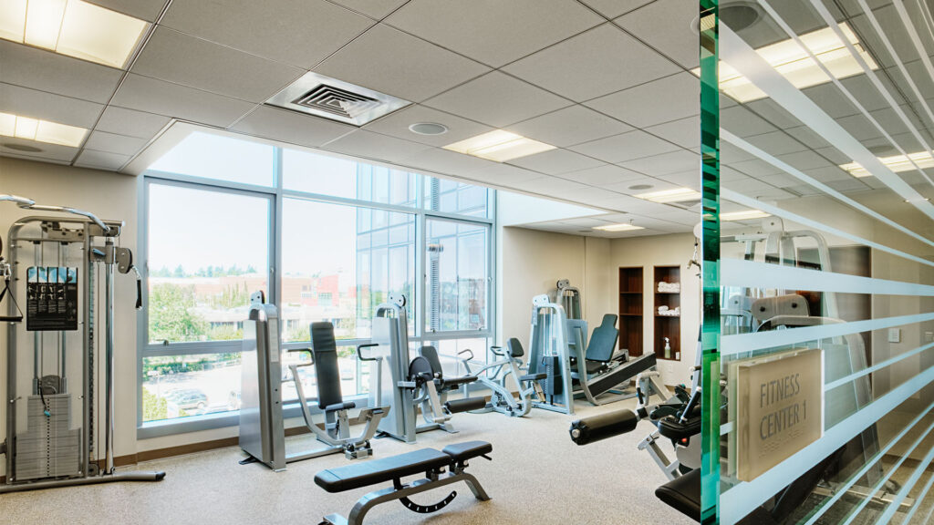 A well-lit gym with various exercise machines, weight benches, and large windows offering an outside view.