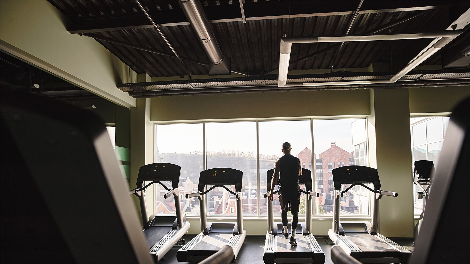 A person stands on a treadmill in a gym, facing large windows with outdoor buildings visible, perhaps pondering workout tips or tricks to enhance their routine.