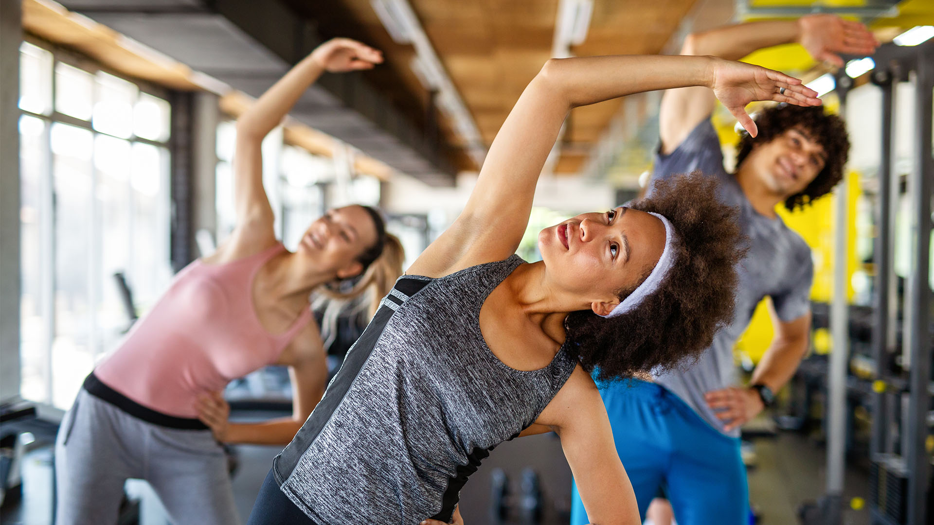 Three people are performing side stretches in a gym, dressed in athletic clothing and facing the camera. It's an ideal snapshot of finding the perfect gym where fitness routines feel both intense and invigorating.