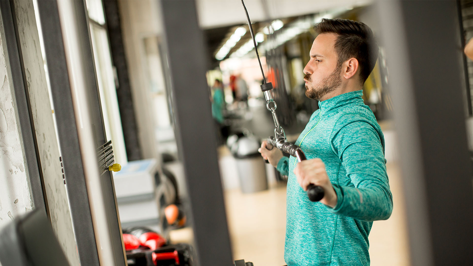 A man wearing a teal long-sleeve shirt exercises on a cable machine at the perfect gym.