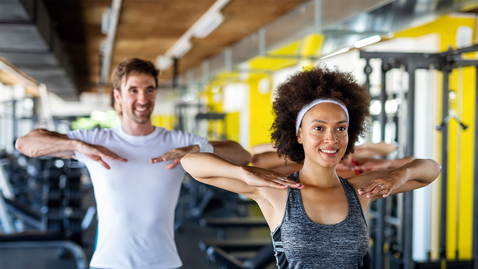 Group of people in workout attire performing exercise moves in a gym with bright yellow walls and fitness equipment in the background, showcasing tips and tricks for finding the perfect gym.