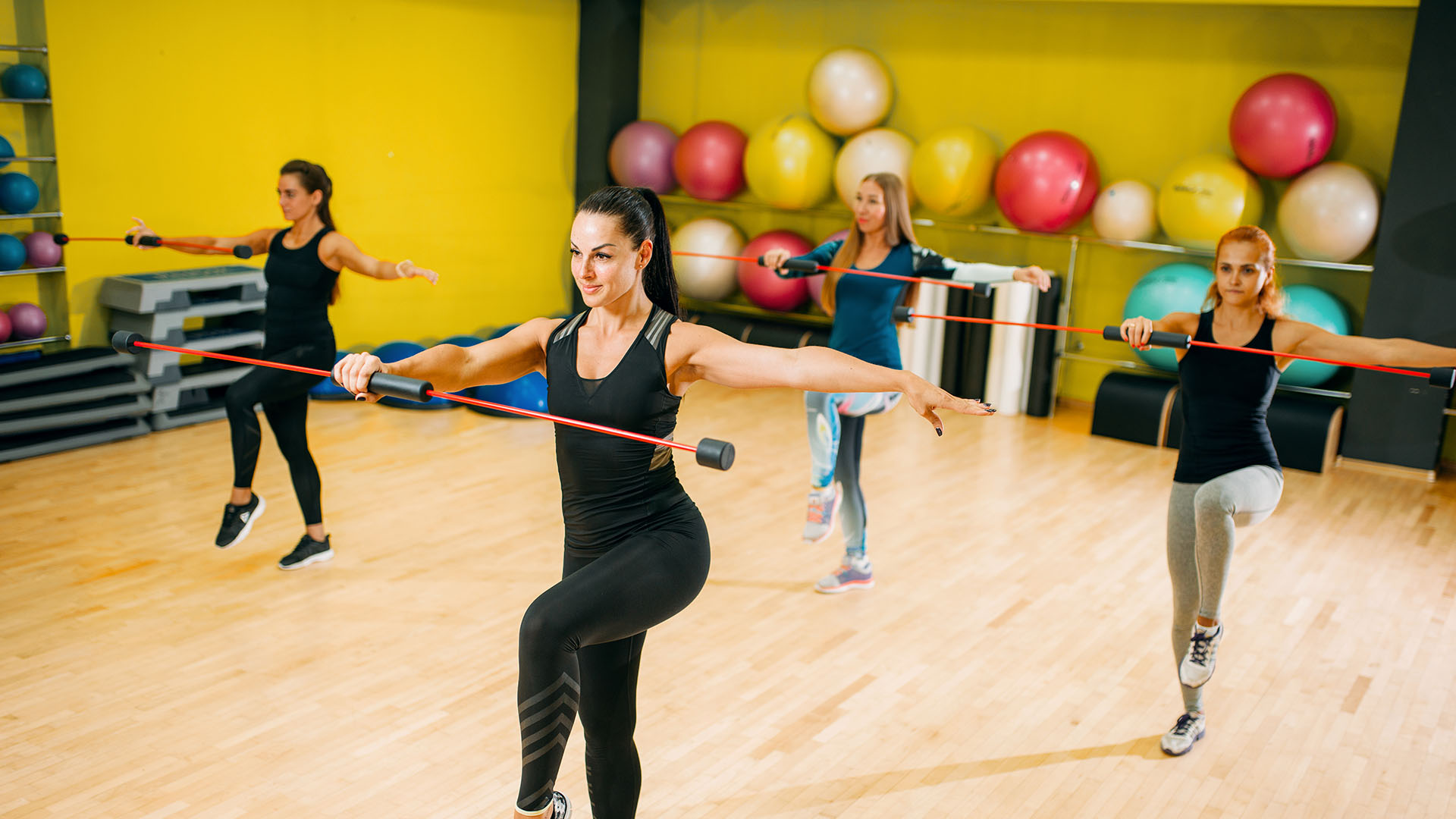 Four women exercising with resistance bars in a bright yellow gym, showcasing how finding the perfect gym can make workouts more enjoyable. Large exercise balls add to the vibrant atmosphere.