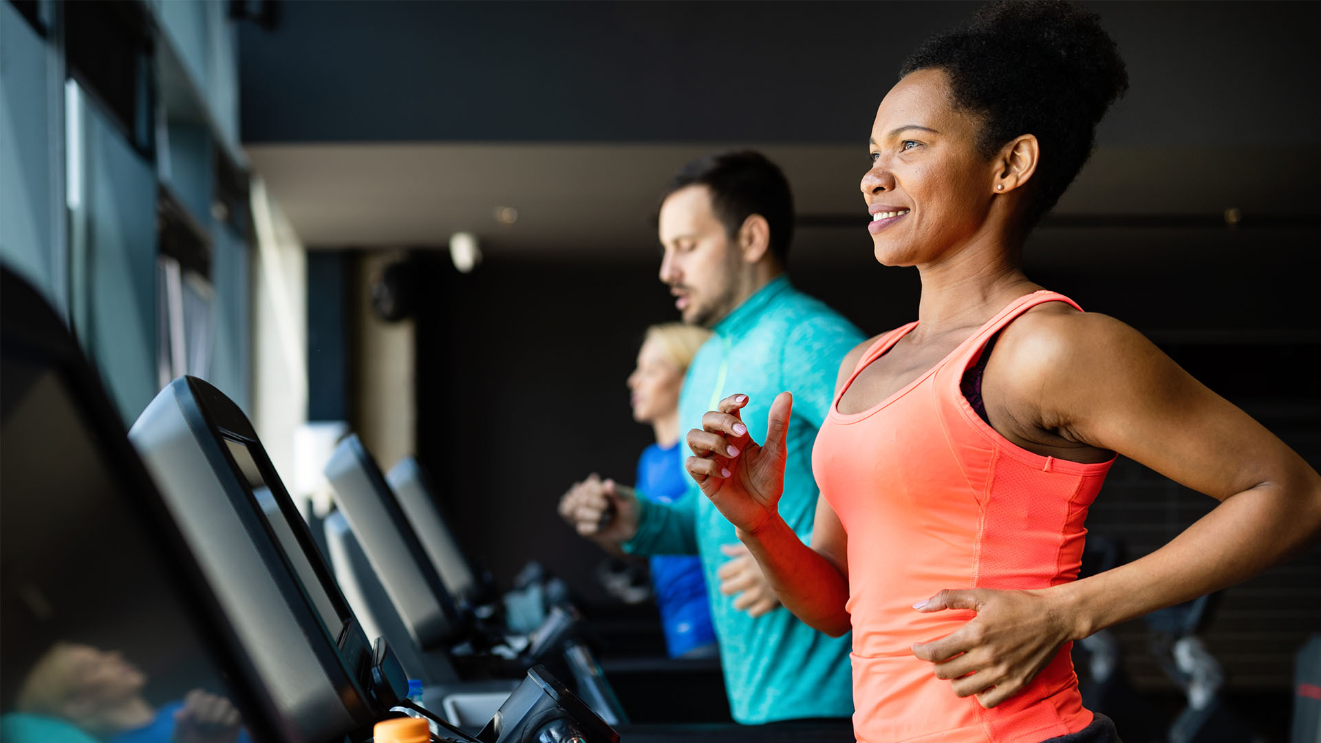 Three people exercise on treadmills in the Perfect Gym. The woman in the foreground, wearing an orange tank top, is smiling.