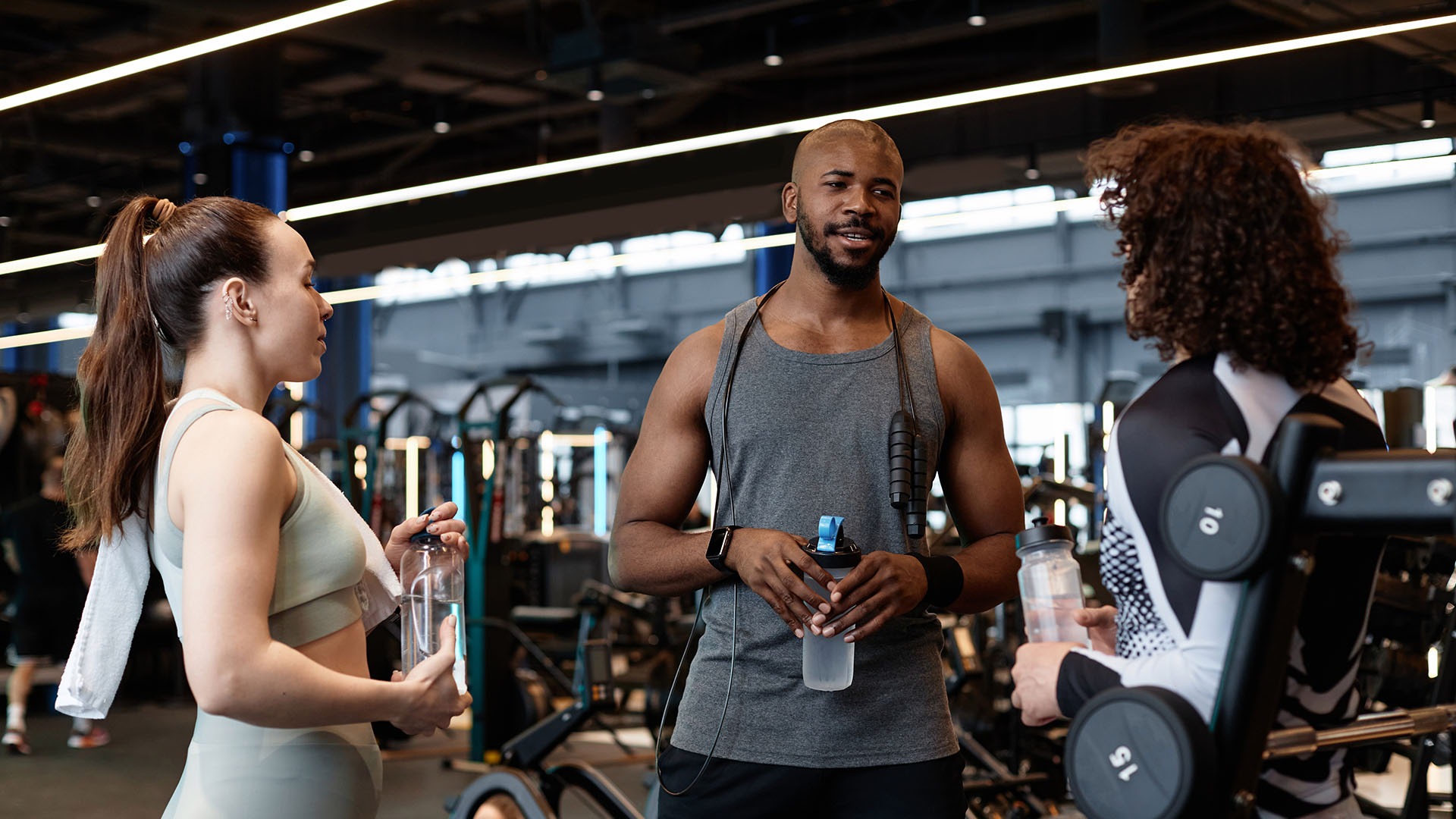 Three people in athletic wear stand and converse at the gym, sharing tips. Two hold water bottles, and various gym equipment is visible in the background.