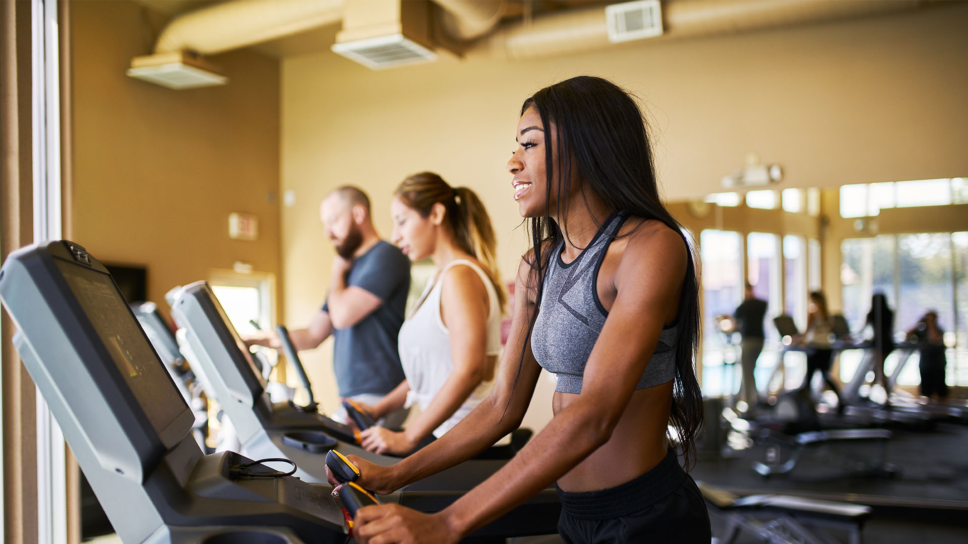 Three people are exercising on treadmills in a gym bathed in natural light. The focus is on the woman in the foreground, wearing a gray sports bra and black leggings, embodying the essence of finding the perfect gym for your fitness journey.