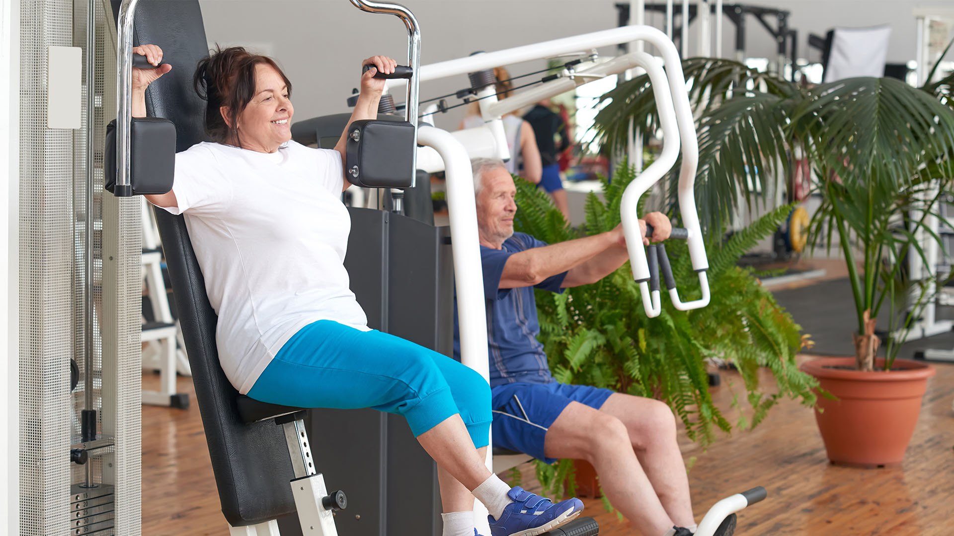 Two people are using exercise machines in a gym. The woman in the foreground, wearing a white shirt and blue pants, seems focused. The man in the background is clad in a blue outfit, both immersed in their routines—just another day at the perfect gym.